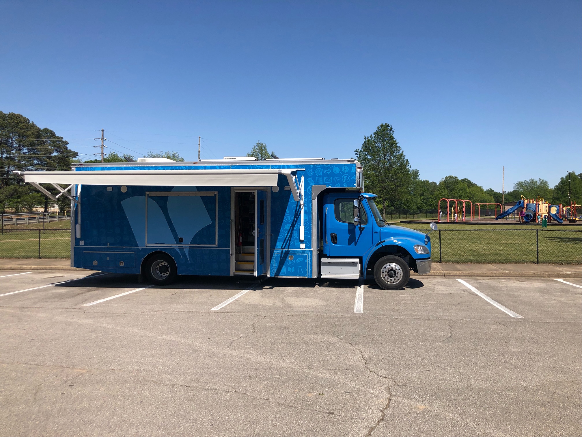 An image of the Bookmobile, a large blue mobile library bus, in a parking lot with its awning extended