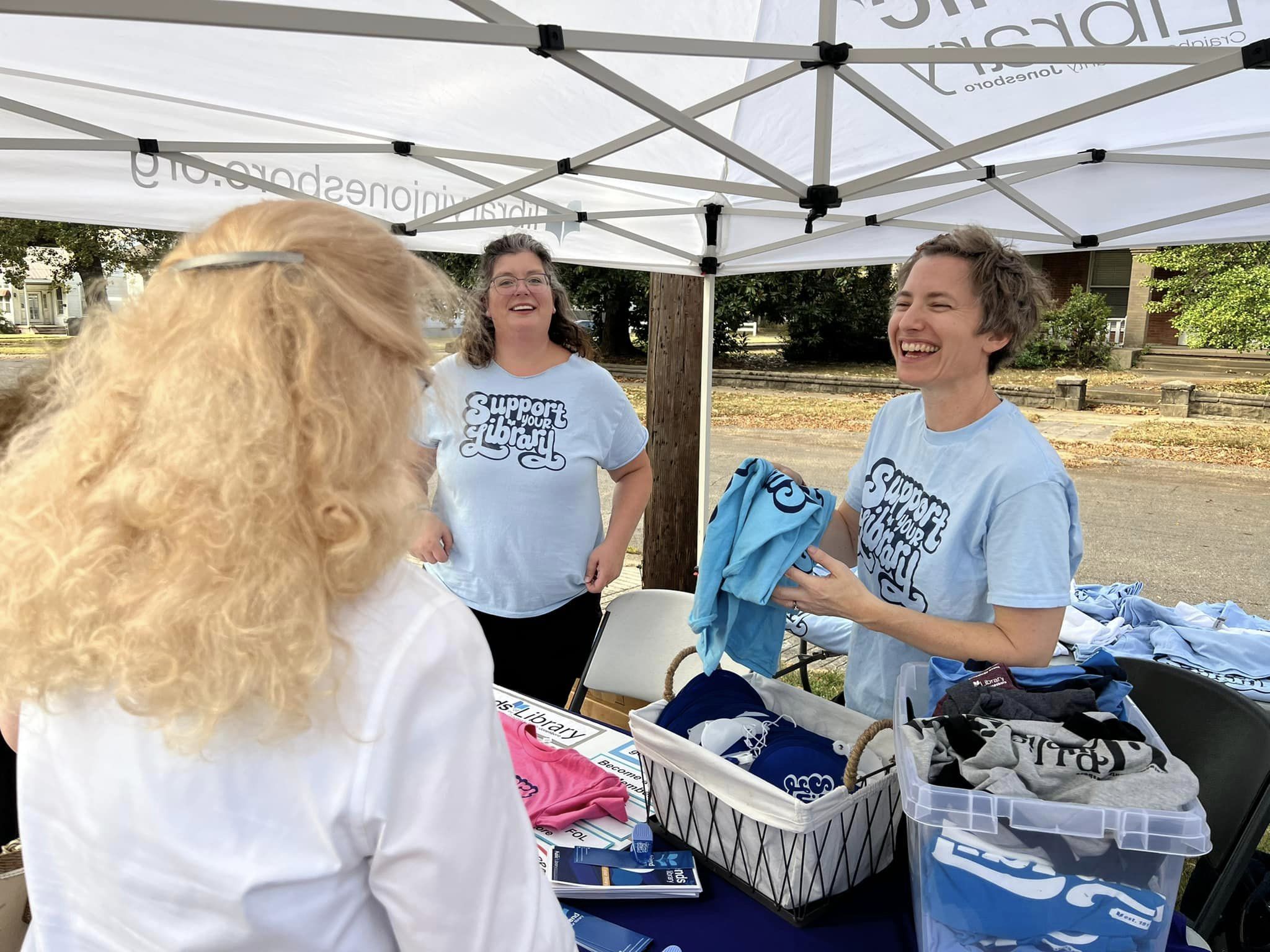Two women pass out shirts at an outdoor Support Your Library event