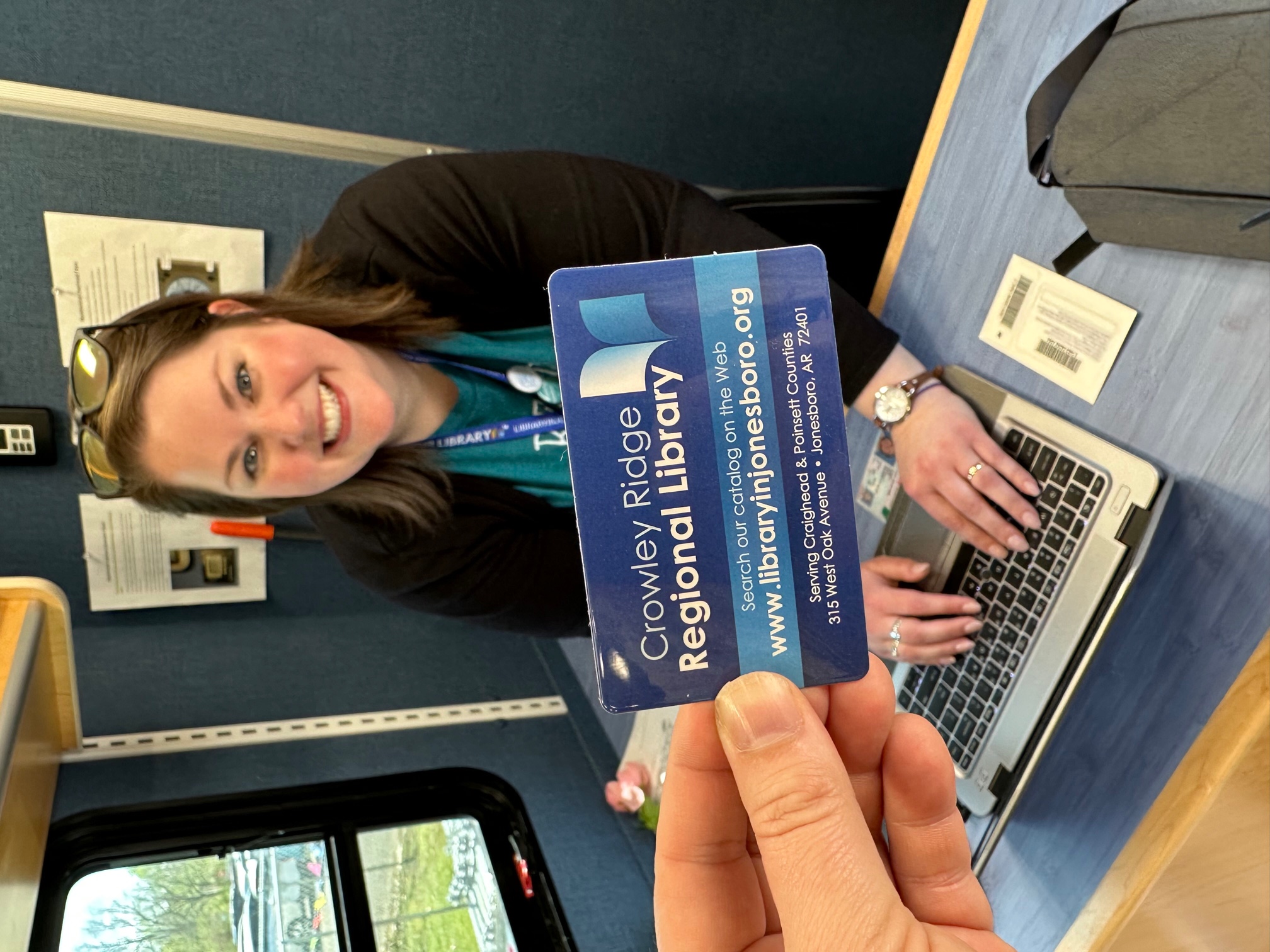 A photo of Ms. Laura, the bookmobile driver, posing with a laptop in front of a hand holding a CCJPL library card